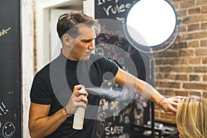 Professional male hairdresser spraying and styling with hairspray his female client's blonde hair in the