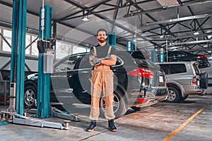 A professional male mechanical employee of a car service workshop stand with crossed arms and tools, looks very symphaticly and