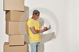 Professional male mover in casual wear making notes, holding clipboard, standing near a stack of cardboard boxes while