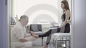 Professional male orthopedist examining young Caucasian woman. Side view wide shot of doctor checking female foot in
