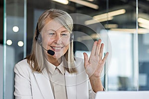 Professional mature woman with headset waving in a friendly manner, representing customer support and positive workplace