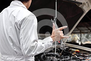 Professional mechanic man holding wrench and his hand with i love you sign with car in open hood at the repair garage.