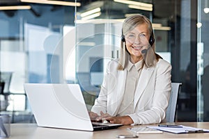 Professional middle-aged woman with headset working on laptop in modern office, displaying customer service and