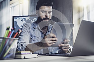 Professional at office desk using tablet in his work.Dark-haired man sitting at window in front of laptop with tablet in his hands