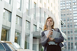 Professional office worker holding coffee, laptop and documents near the business center in city downtown.Thoughtful businesswoman