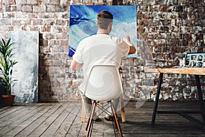 Professional painter sits on chair in front of easel with canvas, hold paintbrush in hand and drawing oil painting. Artist working