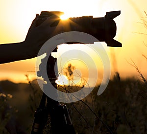 Professional photo camera on a tripod silhouette in the sunset with the lens hood in the hands of women