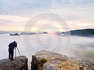 Professional photographer above clouds. Man takes photos with camera on tripod on rocky peak.