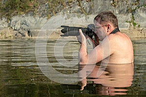 A professional photographer with glasses sits on a mountain river in front of his chest and holds a digital camera in his hands. A