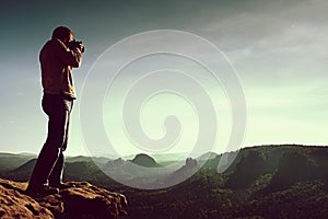 Professional photographer takes photos with mirror camera on cliff of rock. Dreamy misty landscape, hot Sun above