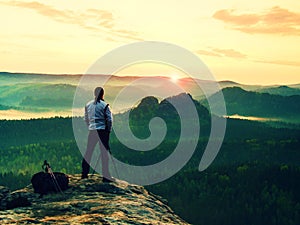 Professional photographer in white jacket takes photos with camera on tripod on rocky peak. Dreamy fogy landscape, spring orange