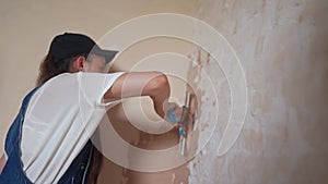 Professional plasterer female spackles the wall, applies and spreads plaster on a spatula in overalls and ball cap