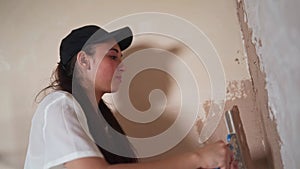 Professional plasterer female spackles the wall, applies and spreads plaster on a spatula in overalls and ball cap