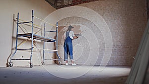 Professional plasterer female spackles the wall, applies and spreads plaster on a spatula in overalls and ball cap