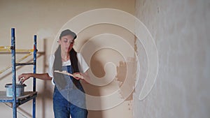 Professional plasterer female spackles the wall, applies and spreads plaster on a spatula in overalls and ball cap