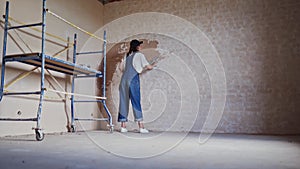 Professional plasterer female spackles the wall, applies and spreads plaster on a spatula in overalls and ball cap