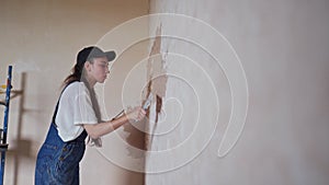 Professional plasterer female spackles the wall, applies and spreads plaster on a spatula in overalls and ball cap