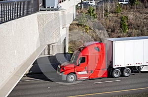 Professional red big rig bonnet semi truck with dry van semi trailer running under the concrete bridge driving on the divided