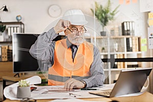 Professional senior male architect builder in white protective helmet and uniform using laptop