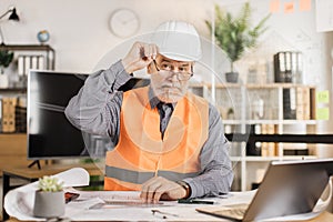 Professional senior male architect builder in white protective helmet and uniform using laptop