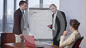 Professional senior manager pointing at graph talking with young colleagues indoors. Portrait of confident serious