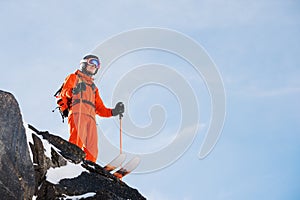 Professional skier-athlete stands on the edge of a high cliff against a blue sky on a sunny day