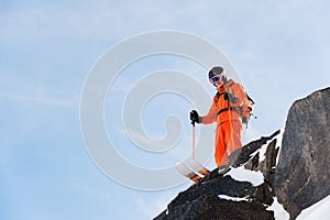 Professional skier-athlete stands on the edge of a high cliff against a blue sky on a sunny day