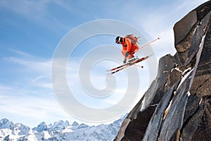 A professional skier makes a jump-drop from a high cliff against a blue sky leaving a trail of snow powder in the