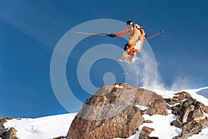 A professional skier makes a jump-drop from a high cliff against a blue sky leaving a trail of snow powder in the