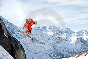 A professional skier makes a jump-drop from a high cliff against a blue sky leaving a trail of snow powder in the