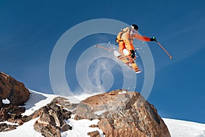 A professional skier makes a jump-drop from a high cliff against a blue sky leaving a trail of snow powder in the