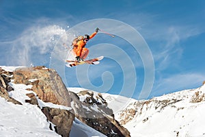 A professional skier makes a jump-drop from a high cliff against a blue sky leaving a trail of snow powder in the