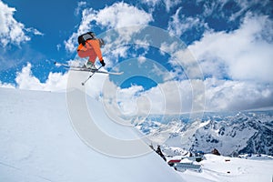 A professional skier in an orange suit jumps from a high cliff against a background of blue sky and clouds, leaving a