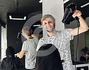 Professional smiling male stylist blow drying woman`s hair with a dryer in salon