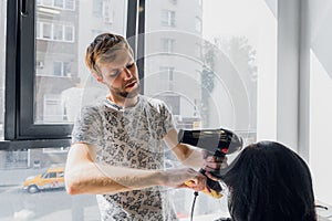 Professional smiling male stylist blow drying woman`s hair with a dryer in salon