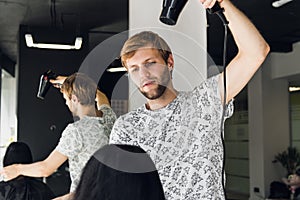 Professional smiling male stylist blow drying woman`s hair with a dryer in salon