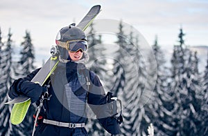 Male skier in goggles with professional ski equipment on his shoulders standing on slope top before skiing in mountains.