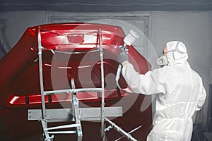 Professional spray painter in protective mask and clothes spraying car hood of vehicle part to red color using spray gun