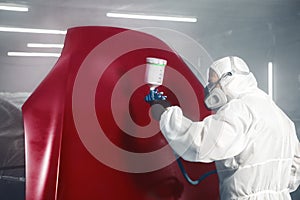 Professional spray painter in protective mask and clothes spraying car hood of vehicle part to red color using spray gun