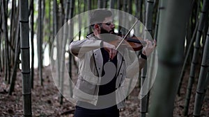 A professional street musician playing the violin plays a composition in a city park standing in the bushes of a bamboo