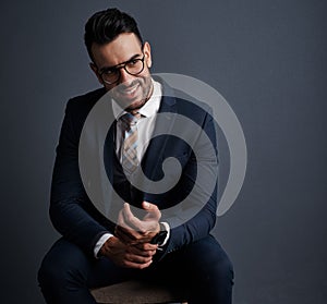 Professional style on point. Studio shot of a stylish young businessman sitting on a chair against a gray background.