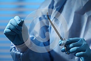 Professional surgeon holding forceps with suture thread on blurred background, closeup. Medical equipment