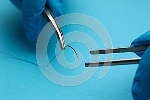 Professional surgeon holding forceps with suture thread on light blue background, closeup. Medical equipment