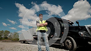 Professional Truck Driver in a yellow waistcoat approaches his truck and crosses his arms Behind Him Parked Long Haul