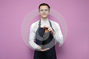 Professional waiter in uniform holds a bottle of red wine and smiles on a pink background, the sommelier recommends drinking wine