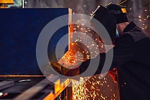 Professional welder wearing protective mask performs work at steel structure manufacturing plant. Yellow sparks spill