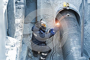 Professional welder welding water or gas steel pipeline in protective trench and mask at construction site pit. City