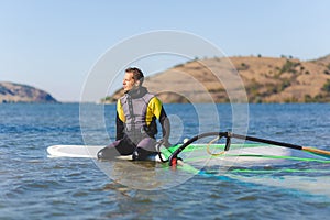 Professional windsurfer wearing wetsuit sitting on board and waiting for wind