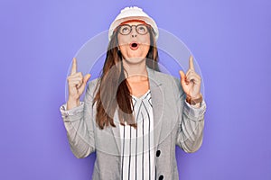 Professional woman engineer wearing industrial safety helmet over pruple background amazed and surprised looking up and pointing
