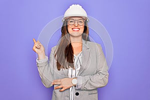 Professional woman engineer wearing industrial safety helmet over pruple background with a big smile on face, pointing with hand